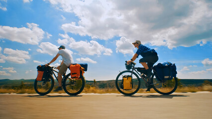 Two cyclists ride their bikes down a scenic road, each with a backpack and panniers on their bikes. A long shot captures their journey across a picturesque countryside. © wkproduction