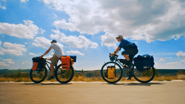 Two cyclists ride their bikes down a scenic road, each with a backpack and panniers on their bikes. A long shot captures their journey across a picturesque countryside.