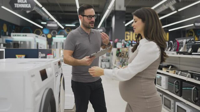 Smiling salesman holding a tablet, discussing and showing washing machine to a pregnant customer, selecting the best options in a modern home appliance store