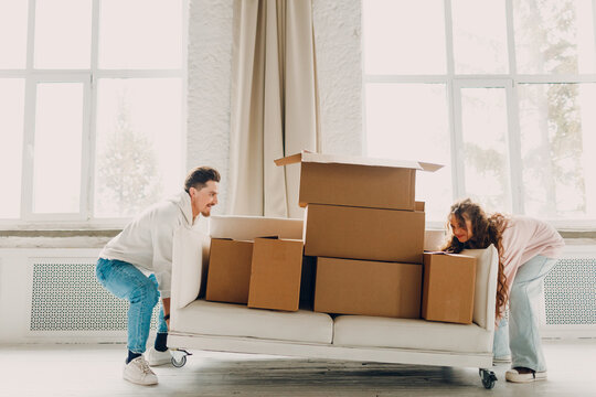 Happy young family couple man and woman push sofa with cardboard boxes in new home. Moving delivery concept.
