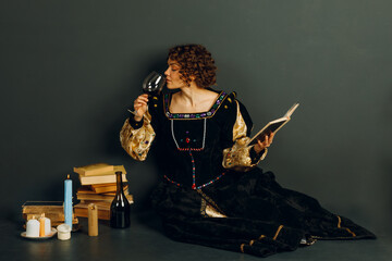 Young adult woman dressed in a medieval dress sitting near ancient books and candles with bottle of red wine and drinking a glass of wine