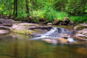 In a lush green forest, a beautiful stream cascades gracefully over the rocks, forming a serene...