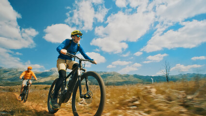 Two cyclists, one in a blue shirt and the other in an orange shirt, ride mountain bikes on a scenic trail. The camera captures a close-up shot of the cyclists.
