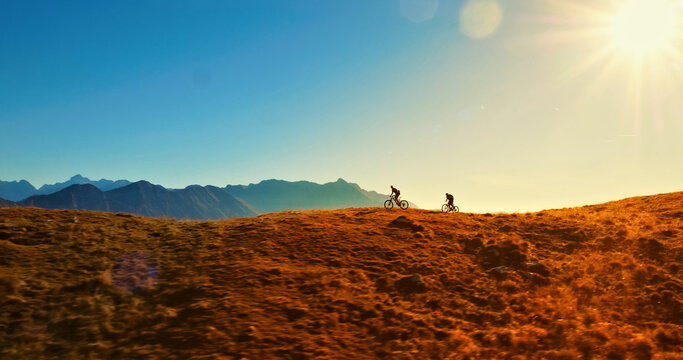 Two mountain bikers traverse a vast, grassy hilltop as the sun sets, casting a warm glow over the landscape. The aerial view highlights the thrill of outdoor adventure and the beauty of nature.