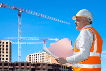 Head of construction company with blueprints. Man architect near houses under construction. Portrait of builder in reflective vest. Man head of construction company watches erection of skyscrapers.