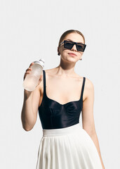 A stylish woman in glasses and fashionable clothes
poses with a water bottle in her hand on a light background