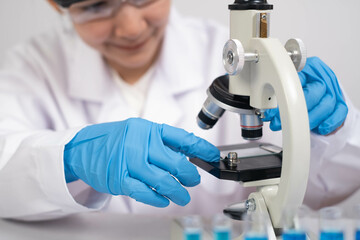 A scientist in a lab coat uses a microscope while wearing blue gloves, focusing on a sample slide,...