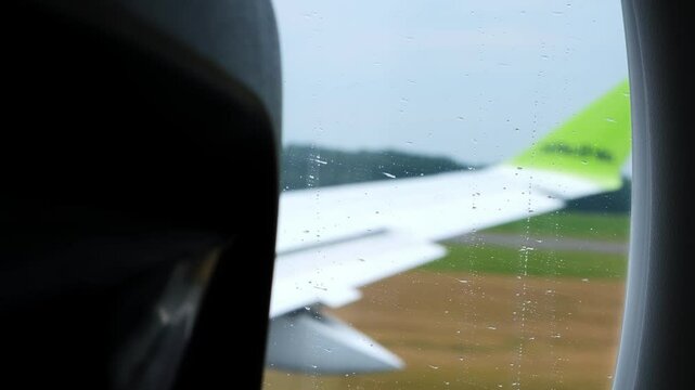 The plane picks up speed on the runway and takes off, lifting off from the ground. Raindrops are running down the glass. View the porthole to the wing of moving passenger airplane. HQ 4K footage.