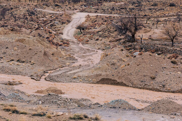 A dry, rugged landscape with a winding dirt road cutting through sparse vegetation, eroded soil,...