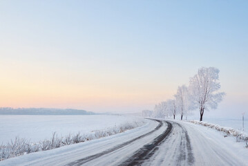 A snowy road winds through a frost-covered landscape at sunrise, with pastel skies and a line of icy trees along a vast winter field.