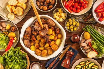 Top view of dinner table with roasted meat and potatoes in ceramic dish surrounded by fresh vegetables mushrooms cheese olives and bread on wooden surface