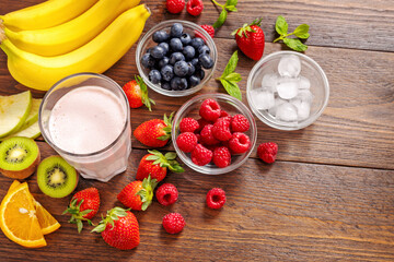 Glass of fresh smoothie with bananas, raspberries, blueberries, strawberries, kiwi and ice cubes on rustic wooden background. Healthy ingredients for cooking summer drink