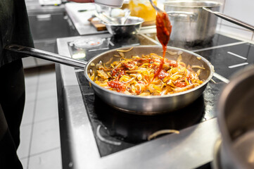 A chef prepares a dish in a stainless steel pan on a stovetop, adding tomato sauce to sautéed...
