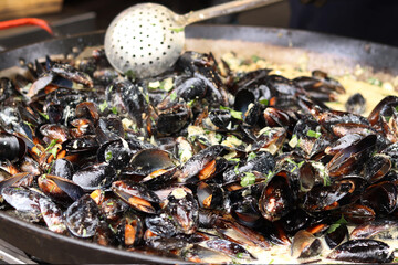 Mussels with parsley and sauce in a large frying pan. Food court. Street food. Mussels with shells cooked with herbs in a frying pan. Mussels cooked in sauce at a street market. Cooking a seafood dish