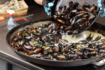 Mussels with parsley and sauce in a large frying pan. Food court. Street food. Mussels with shells cooked with herbs in a frying pan. Mussels cooked in sauce at a street market. Cooking a seafood dish