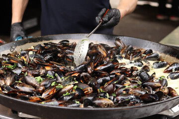 Mussels with parsley and sauce in a large frying pan. Food court. Street food. Mussels with shells cooked with herbs in a frying pan. Mussels cooked in sauce at a street market. Cooking a seafood dish