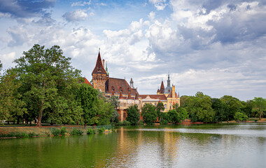 Obraz premium Vajdahunyad Castle stands majestically by a peaceful lake, surrounded by lush greenery and dramatic clouds in Budapest, Hungary.