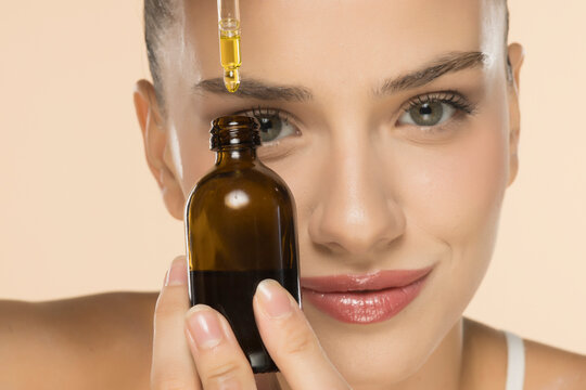 Young woman demonstrating skincare serum application from amber glass dropper bottle on forehead, smiling on beige background.