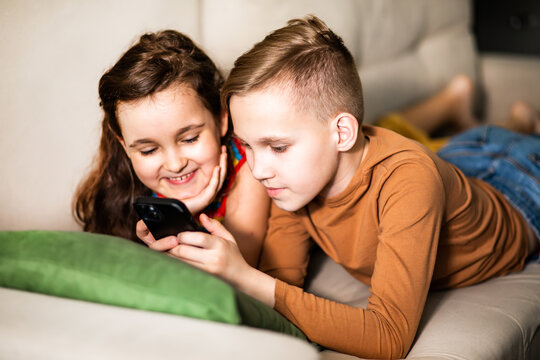 Cute little girl and boy teenager using smart phone lies on sofa in living room at home together.