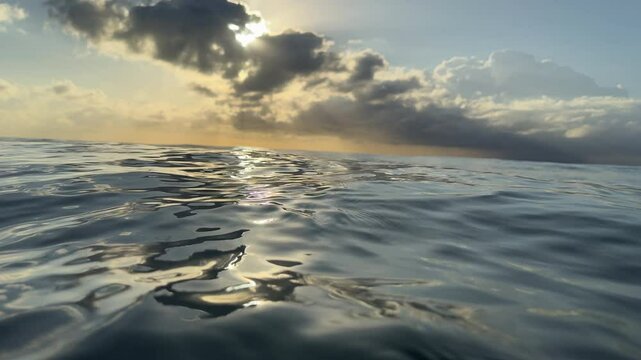 A golden sun rising over the ocean at dawn with rays beaming out from puffy clouds colouring the ocean with beautiful silvery tones. Captured in slow motion within the ocean on sunrise. 