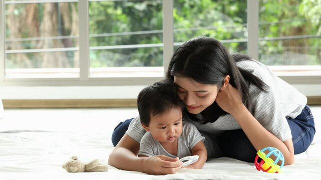 Young Asian mother lovingly embraces baby from behind and lie on soft white blanket while toddler holds teether in living room near large window