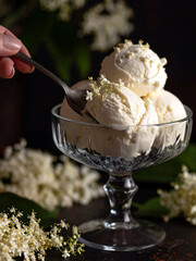 A hand scoops ice cream into a bowl, next to which white elderflowers are elegantly arranged.