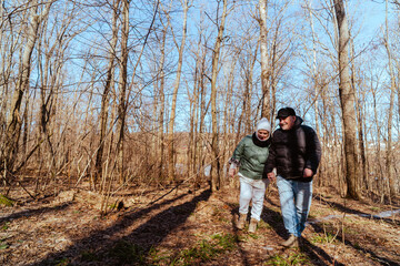 Obraz premium An elderly couple, a man and a woman, walk through a spring forest, enjoying the beauty of nature. Sunlight filters through fresh green leaves, creating a serene and romantic atmosphere