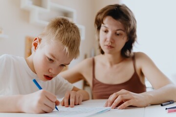 A loving mother and her son work together on homework at the kitchen table, symbolizing family...