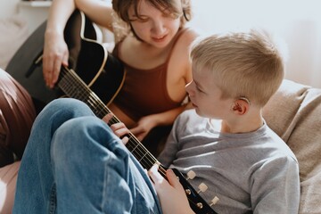 Mother and son playing guitar together, family love. The child wears a hearing aid, highlighting...