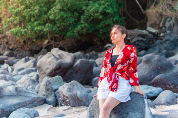 An Indonesian woman sits barefoot on a rock at the beach, wearing a floral red top and white...
