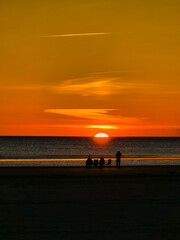 Fototapeta premium Silhouetted figures gather along the beach in Denmark as the sun sets on the horizon, casting warm orange and yellow hues across the sky and reflecting on the calm waters.