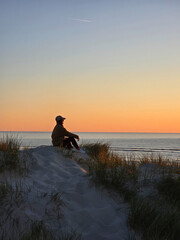 A person sits atop a sandy dune, gazing at the colorful sunset over the calm waters of the Danish...