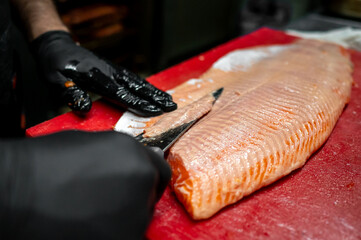 A person wearing black gloves fillets fresh salmon on a red cutting board using a sharp knife,...