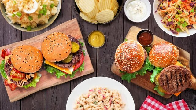 BBQ table scene with hamburgers, classic salads and snacks. Stop motion table setting placement video. Top view over a dark wood background.