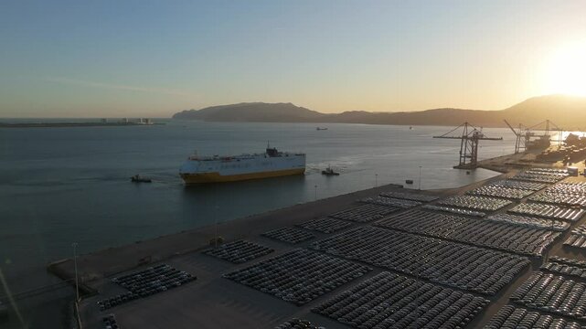 Ro-Ro Cargo Ship with Tugboats Arriving and New Cars on Large Full Parking Lot at Sunset in Port. Car Storage, Automotive Industry. Ocean and Mountains. Setubal, Portugal. Aerial View. Orbiting