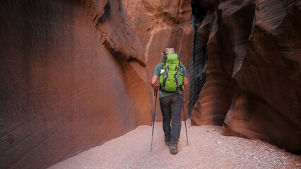Adult man hiker with backpack and trekking poles hiking on bottom dry sandy cave riverbed in deep slot canyon with curved and smooth sandstone rocks walls of orange red color, slow motion, view back