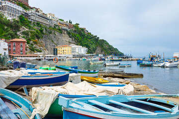 Colorful wooden fishing boats on shore and moored in Marina Grande harbor, Sorrento, Campania, Italy. Coastal buildings on cliffs and calm Tyrrhenian Sea under overcast sky.