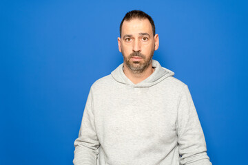 Hispanic man with a beard in his 40s wearing a gray sweatshirt looks straight ahead with a serious and worried expression, isolated on a blue studio background.