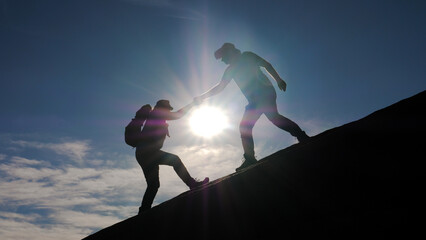 Teamwork two tourists helps to travel. Silhouette helping hand between two climbers rising to top of mountain. People hikers help when climbing mountain or overcoming difficulties for victory success