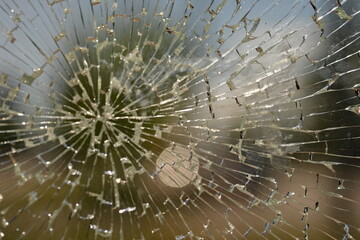 shattered safety glass on a public transport bus stop. The cracks form a circular spiderweb pattern, creating an abstract texture with sharp reflections. The background is softly blurred and shows tre