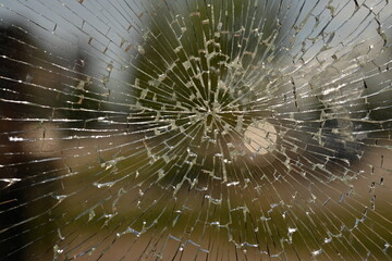 shattered safety glass on a public transport bus stop. The cracks form a circular spiderweb pattern, creating an abstract texture with sharp reflections. The background is softly blurred and shows tre