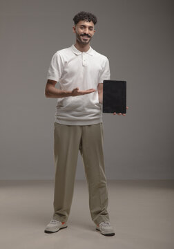 A young Saudi man in modern casual attire holding a blank white foam board for advertisement or mockup purposes, standing in a studio with a neutral background. Ideal for marketing use.