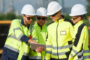 Engineering team in safety gear planning pipeline maintenance at oil and gas refinery site with storage tanks in the background.