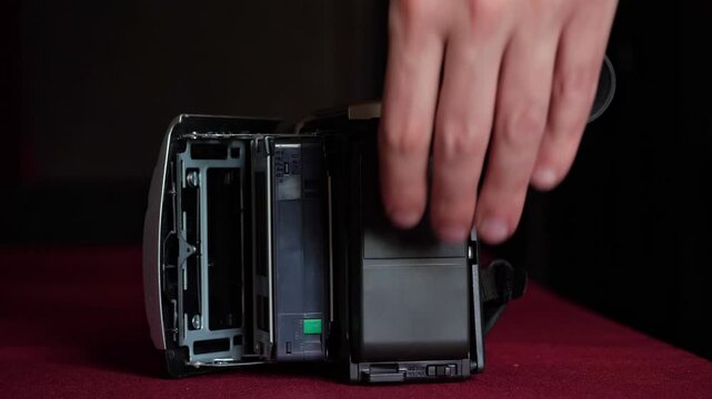 A close-up shot of a hand inserting an S-VHS video cassette into the side loading mechanism of a vintage video camera. The black cassette and the camera's textured body are clearly visible