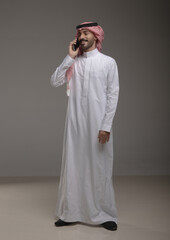 Saudi Arabian man wearing traditional thobe and shemagh using a smartphone, making a call and...
