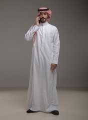 Saudi Arabian man wearing traditional thobe and shemagh using a smartphone, making a call and...