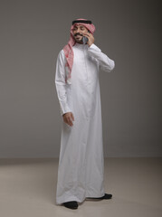 Saudi Arabian man wearing traditional thobe and shemagh using a smartphone, making a call and...