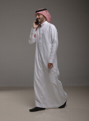 Saudi Arabian man wearing traditional thobe and shemagh using a smartphone, making a call and...