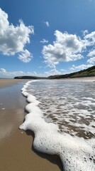 Gentle ocean waves roll steadily over a sandy beach beneath a radiant, sunny sky. Vertical.