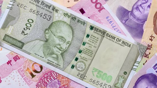 Indian rupee banknote falling in slow motion on Chinese Yuan banknotes background. Concept of international trade restrictions, tariff wars for economic dominance, emerging markets of India and China.